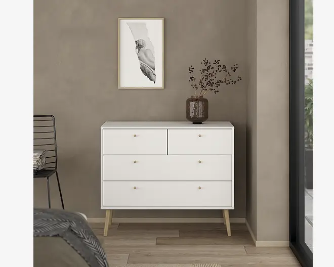 Et moderne, minimalistisk skab med mat hvid finish, der har fire skuffer med små runde guldfarvede greb. Skabet står på skråtstillede ben af lyst træ, hvilket giver det et retro udtryk. På toppen står en mørk keramisk vase med tørrede grene. Det samlede design er enkelt og stilrent, velegnet til moderne indretning.