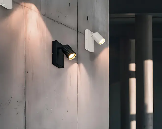 To vægmonterede spotlys vises, det ene i mat sort og det andet i mat hvid. Begge har et cylindrisk, rillet lampehoved, der kan vinkles for at rette lyset, og er monteret på en rektangulær base. Designet er minimalistisk og moderne, velegnet til accent- eller arbejdsbelysning. Lamperne kaster fokuserede lysstråler på væggen, hvilket fremhæver deres justerbarhed og lysets kvalitet.
