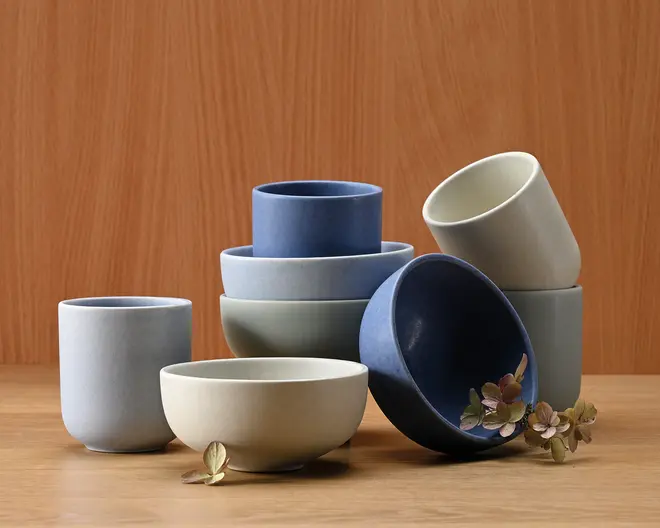 Et sæt minimalistiske keramikskåle og -kopper i afdæmpede nuancer af blå, grå og offwhite. Delene har en glat, mat overflade og enkle, rene linjer, hvilket giver dem et moderne og elegant udtryk. Skålene og kopperne kan stables, og nogle er vist stablet sammen. Et par tørrede blomster er placeret omkring sættet som dekoration.