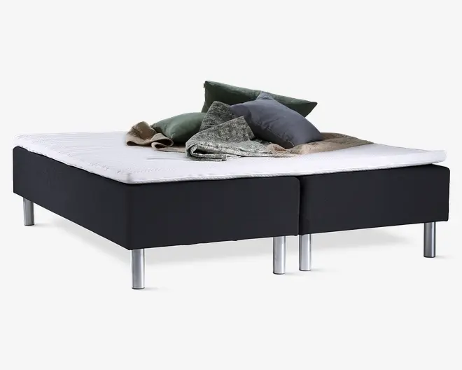 En moderne seng med sort polstret bund og sølvfarvede metalben. Madrassen er hvid og har en mellemtyk profil. Ovenpå ligger der flere pyntepuder i grønne og grå nuancer samt et tekstureret gråt tæppe og et beige tæppe, hvilket giver sengen et hyggeligt og indbydende udtryk.