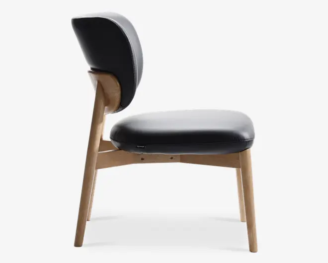 Sidebillede af en stol med en tyk, polstret sædehynde og rygl sopol parret i glat sort materiale. Stel extra ben er lavet af lyst træ med en naturlige finish og har et let vinklet, retro-inspireret design farm 1950'erne. Ryglæ awhile er let buet for ergonomisk støtte, og sædet size bredt og komfortabelt ud.