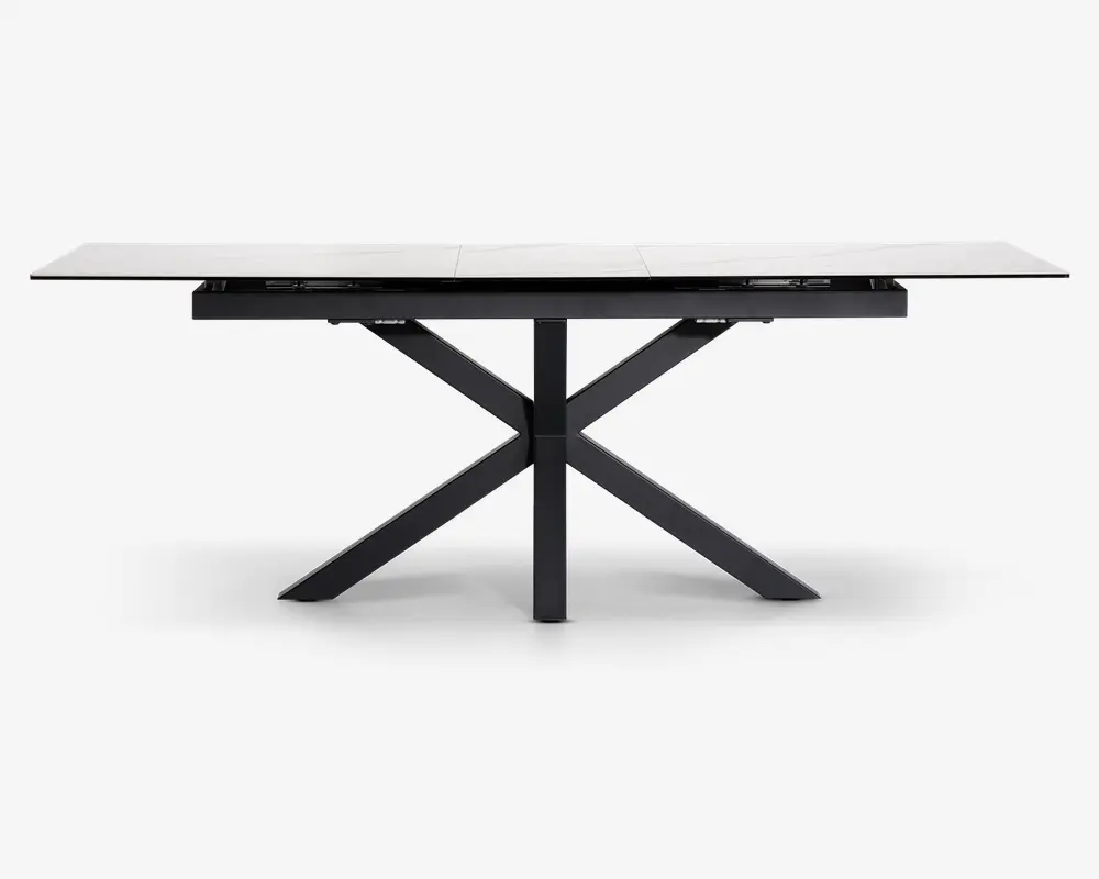 Rektangulært bord med en hvid, marmorlignende bordplade og et moderne sort metalstel. Bordpladen ser ud til at være udtrækkelig med synlige samlinger, der indikerer tillægsplader. Stellet har et geometrisk krydsben-design, som giver stabilitet og et moderne udtryk.