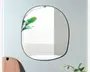 Vægmonteret spejl med en uregelmæssig, afrundet form og en tynd sort ramme. Spejlet har et moderne, minimalistisk design og reflekterer et lyst rum med et vindue med vandrette persienner. Dekorative elementer som en trægenstand og grønne plantegrene er delvist synlige i forgrunden.