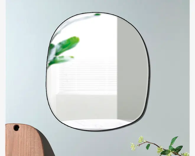 Vægmonteret spejl med en uregelmæssig, afrundet form og en tynd sort ramme. Spejlet har et moderne, minimalistisk design og reflekterer et lyst rum med et vindue med vandrette persienner. Dekorative elementer som en trægenstand og grønne plantegrene er delvist synlige i forgrunden.