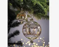 To klare glaskugler hænger fra en gren på et juletræ. Kuglerne er kugleformede og har detaljerede guldfarvede dekorationer, herunder elegante linjer, stjernemotiver og geometriske mønstre. Glasset er blankt og giver et eksklusivt og reflekterende udtryk. Kuglerne er ophængt i tynde guldsnore, og baggrunden er blødt sløret med varme, festlige lys.