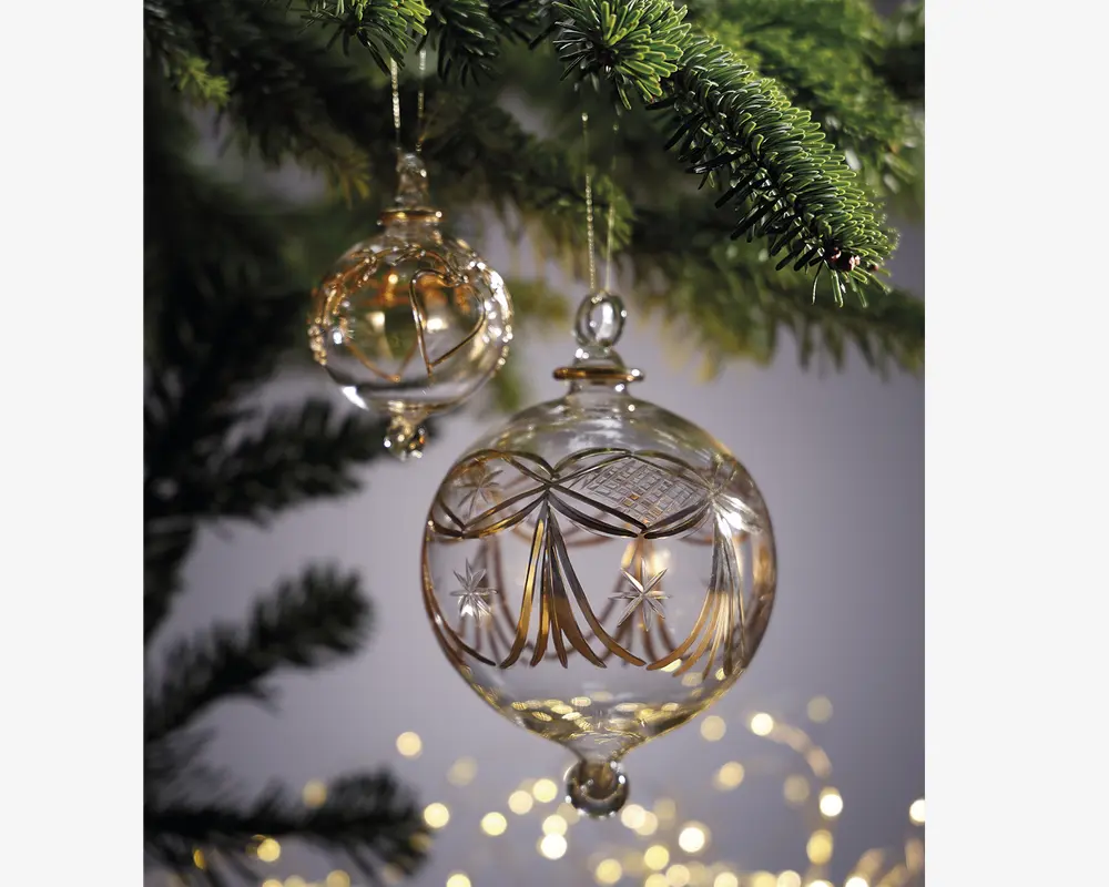 To klare glaskugler hænger fra en gren på et juletræ. Kuglerne er kugleformede og har detaljerede guldfarvede dekorationer, herunder elegante linjer, stjernemotiver og geometriske mønstre. Glasset er blankt og giver et eksklusivt og reflekterende udtryk. Kuglerne er ophængt i tynde guldsnore, og baggrunden er blødt sløret med varme, festlige lys.