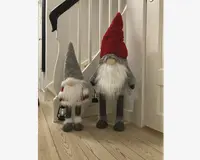 To plyssede nissefigurer med lange, hvide skæg og runde næser. Den største nisse har en høj, spids rød hue, grå krop og lange ben, mens den mindre nisse har en grå hue, rød-hvide vanter og holder en lille sort lanterne. Begge har bløde, lodne teksturer og er designet til at stå oprejst.