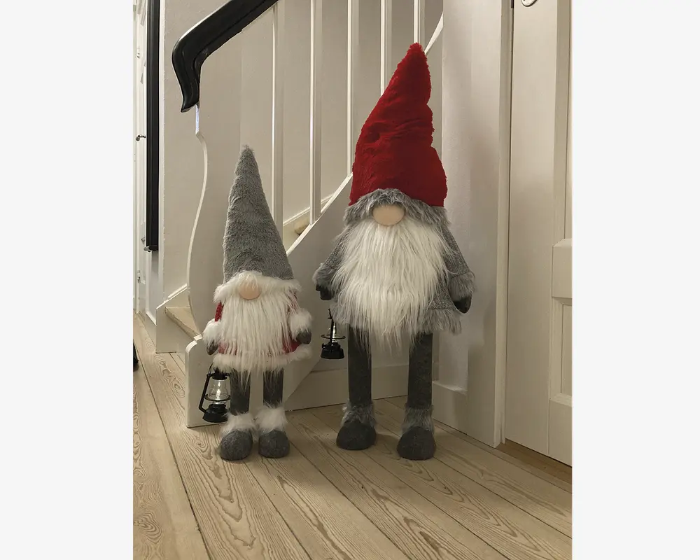 To plyssede nissefigurer med lange, hvide skæg og runde næser. Den største nisse har en høj, spids rød hue, grå krop og lange ben, mens den mindre nisse har en grå hue, rød-hvide vanter og holder en lille sort lanterne. Begge har bløde, lodne teksturer og er designet til at stå oprejst.