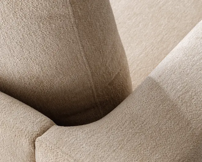 Nærbillede af en beige polstret overflade med tekstureret, boucle-lignende stof. Billedet fremhæver det bløde, fyldige materiale og den buede, polstrede struktur, med tydelige syninger langs sømmene. Stoffet virker slidstærkt og komfortabelt, egnet til daglig brug.