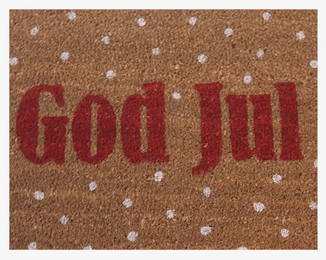 Brun kokosmåtte med teksten 'God Jul' i store, røde bogstaver midt på. Overfladen er dekoreret med jævnt fordelte hvide prikker, der ligner snefnug og giver måtten et julet og festligt udtryk.