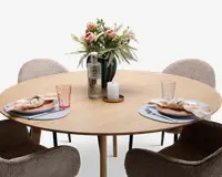 Et rundt bord i lyst træ dækket til to personer med to beige polstrede stole. Hver kuvert har en rund dækkeserviet, en tallerken med en foldet stofserviet, en gaffel og kniv samt et klart drikkeglas med et let rosa skær. Bordet er pyntet med en høj vase med lyserøde og hvide blomster, en mønstret glasflaske og et hvidt bloklys på en træbakke.