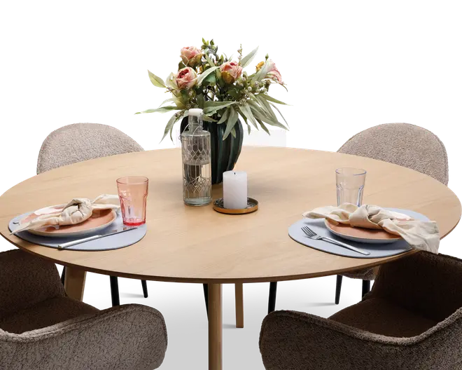 Et rundt bord i lyst træ dækket til to personer med to beige polstrede stole. Hver kuvert har en rund dækkeserviet, en tallerken med en foldet stofserviet, en gaffel og kniv samt et klart drikkeglas med et let rosa skær. Bordet er pyntet med en høj vase med lyserøde og hvide blomster, en mønstret glasflaske og et hvidt bloklys på en træbakke.