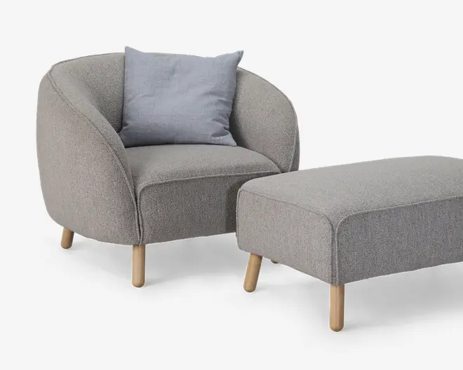 Et moderne lænestol- og fodskammelsæt betrukket med lysegråt stof. Lænestolen har en rundet ryg og sider, en tyk polstret sædehynde og fire lyse træben. Der medfølger en enkelt lyseblå-grå pyntepude for ekstra komfort. Fodskamlen er rektangulær med afrundede kanter og står også på fire lyse træben, som matcher stolens design.