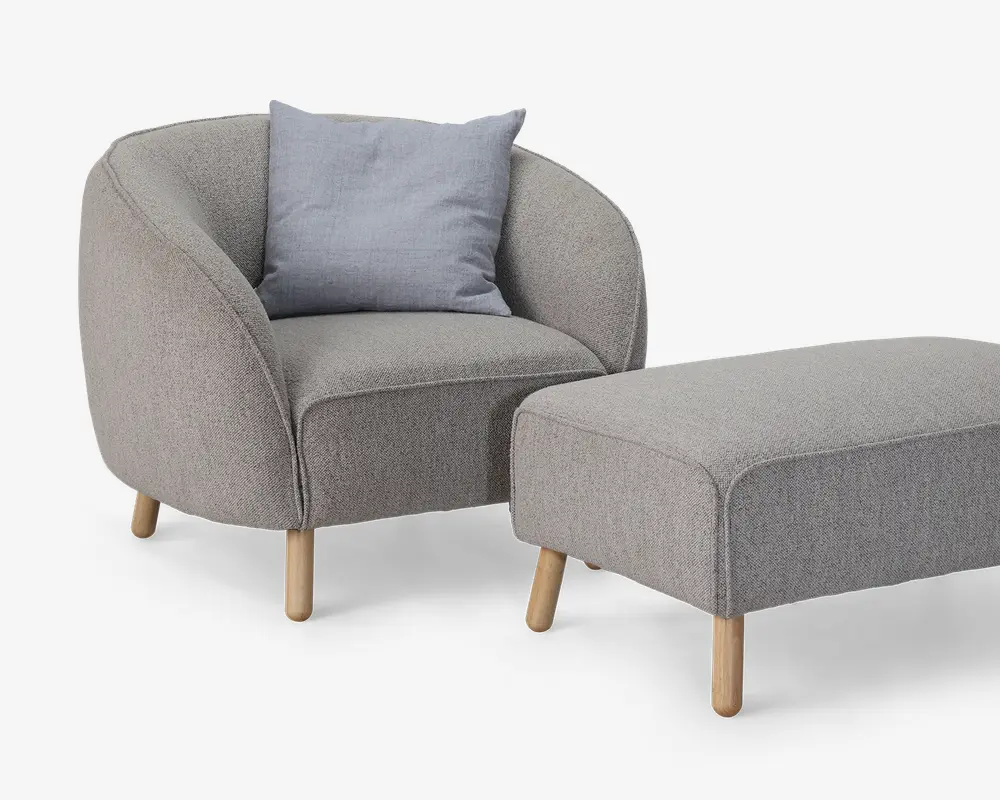 Et moderne lænestol- og fodskammelsæt betrukket med lysegråt stof. Lænestolen har en rundet ryg og sider, en tyk polstret sædehynde og fire lyse træben. Der medfølger en enkelt lyseblå-grå pyntepude for ekstra komfort. Fodskamlen er rektangulær med afrundede kanter og står også på fire lyse træben, som matcher stolens design.