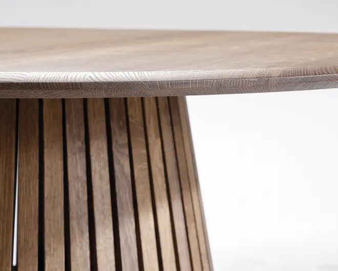 Nærbillede af en træoverflade med en glat, afrundet kant og en base bestående af lodrette trælameller, der er arrangeret i et bredt, geometrisk mønster. Træets årer er synlige og giver et naturligt og tekstureret udtryk.