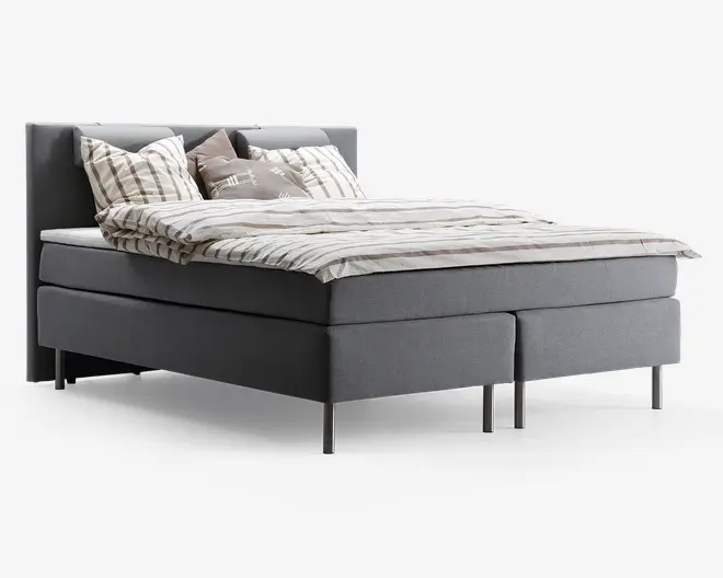 En moderne seng med gråt polstret stel og sengegavl, understøttet af slanke metalben. Sengen er redt med et lyst, stribet dynebetræk og flere puder i forskellige neutrale nuancer og mønstre, hvilket giver et hyggeligt og indbydende udtryk.