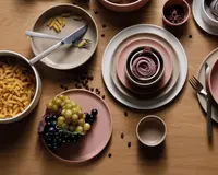 Et koordineret sæt keramikservice i afdæmpede rosa og beige nuancer, bestående af tallerkener, skåle og kopper. Sættet har et moderne, minimalistisk design med glatte, matte overflader og afrundede kanter. Tallerkener og skåle er stablet pænt, og sættet vises med bestik i rustfrit stål. Service sættet er præsenteret med madvarer som druer, pasta og kaffebønner, hvilket fremhæver dets alsidighed til forskellige måltider.