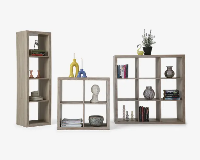 Tre reoler i lyst træ i forskellige størrelser og udformninger vises. Hylderne har en naturlig træstruktur og åbne rum i kubestil. De er dekoreret med forskellige genstande såsom bøger, lys, vaser, en potteplante og dekorative objekter. Reolerne er alsidige og kan bruges både lodret og vandret, hvilket giver fleksible opbevarings- og udstillingsmuligheder.