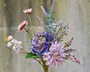 En buket med forskellige kunstige blomster i nuancer af lilla, pink og lavendel. Arrangementet indeholder store, naturtro blomster med lagdelte kronblade, mindre accentblomster og grønne blade. Stilkene er synlige, og blomsterne har en realistisk tekstur og farveovergang, hvilket giver et naturligt udseende.