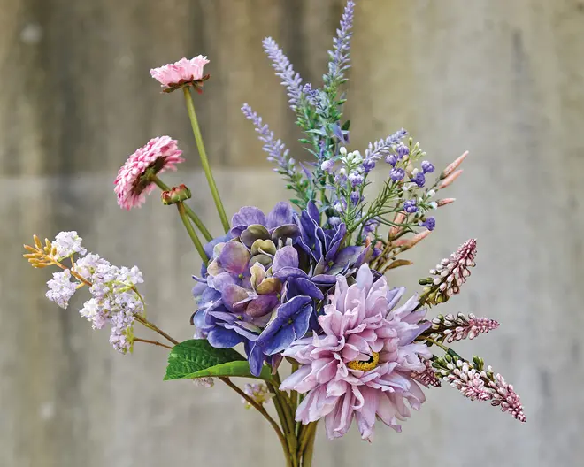 En buket med forskellige kunstige blomster i nuancer af lilla, pink og lavendel. Arrangementet indeholder store, naturtro blomster med lagdelte kronblade, mindre accentblomster og grønne blade. Stilkene er synlige, og blomsterne har en realistisk tekstur og farveovergang, hvilket giver et naturligt udseende.