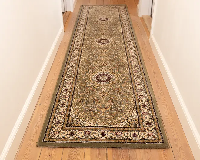 En lang, rektangulær løber med et indviklet blomster- og medaljonmønster i dæmpede jordfarver, herunder olivengrøn, beige og bordeaux. Tæppet har tre cirkulære medaljoner jævnt fordelt langs længden, omgivet af en detaljeret kant med snoede vinmønstre. Kanterne er pænt afsluttet, og tæppet ligger fladt på et lyst trægulv i en gang.