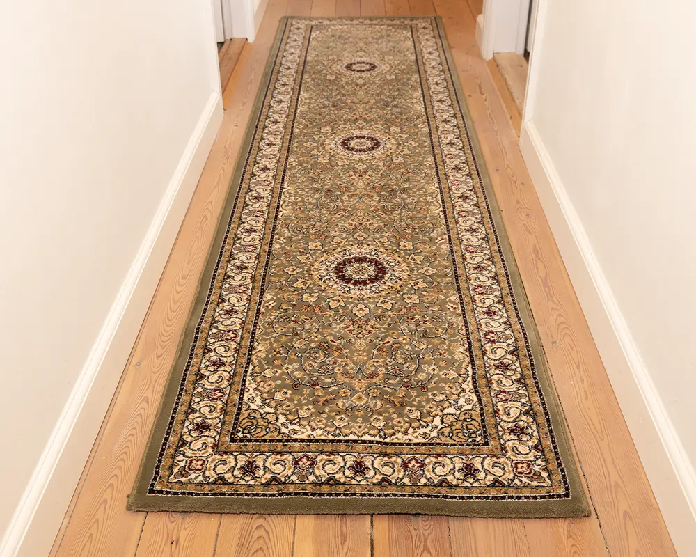 En lang, rektangulær løber med et indviklet blomster- og medaljonmønster i dæmpede jordfarver, herunder olivengrøn, beige og bordeaux. Tæppet har tre cirkulære medaljoner jævnt fordelt langs længden, omgivet af en detaljeret kant med snoede vinmønstre. Kanterne er pænt afsluttet, og tæppet ligger fladt på et lyst trægulv i en gang.