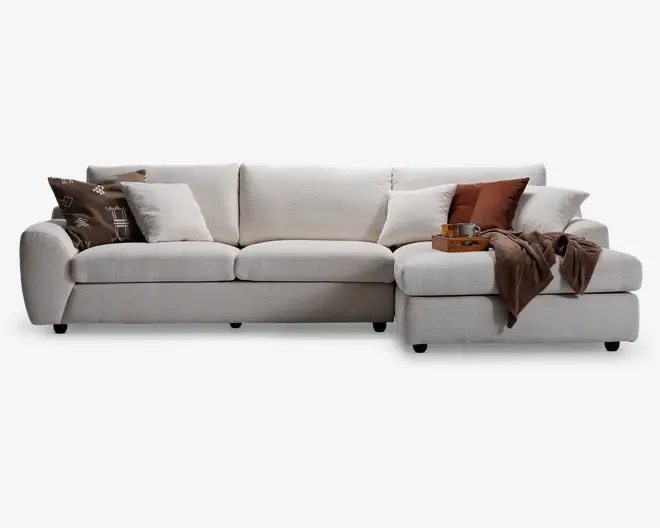 En moderne L-formet sofa med blødt, lysebeige betræk og dybe, polstrede sæder. Sofaen har brede, afrundede armlæn og leveres med flere pyntepuder i forskellige farver og mønstre, herunder brun, rust og creme. Et blødt, brunt tæppe er lagt over chaiselongen, og et træfad med en kop og småting står ovenpå, hvilket giver et hyggeligt og indbydende udtryk.