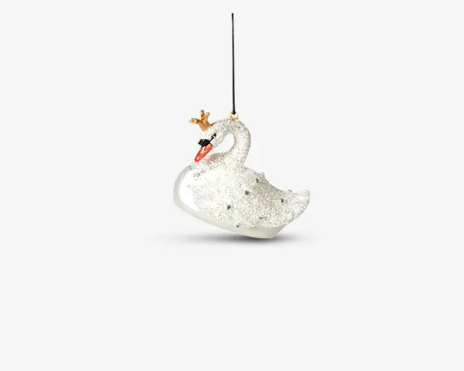 Hvid svaneformet pynt dækket af glitrende glimmer, med en lille guldkron på hovedet og et klart orange næb. Ornamentet er udsmykket med små ædelstenslignende detaljer og hænger i et tyndt sort bånd.