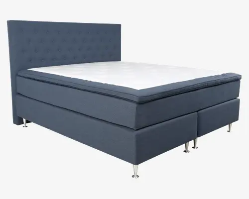 Moderne seng med høj, polstret sengegavl, der har et knap-tuftet design. Sengerammen og bunden er betrukket med mørkeblåt stof, og madrassen har et tykt, blødt toplag for ekstra komfort. Sengen står på slanke, metalliske ben, hvilket giver et moderne udtryk og gør det nemt at rengøre under sengen.