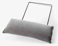 Rektangulær, tekstureret lysegrå pude med et tæt vævet stofbetræk. Puden er fastgjort til en tynd, sort metalramme, som ser ud til at være beregnet til ophængning eller støtte af puden. Pudens kanter er pænt syet, og det samlede design er minimalistisk og moderne.