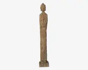 Høj, slank træskulptur med en grov, tekstureret overflade og rektangulær sokkel. Figuren har forlængede proportioner, med arme udskåret tæt ind til kroppen og minimal detaljering på overkrop og lemmer. Hovedet er afrundet og glat med en lille topknude, og ansigtstrækkene er slørede. Udtrykket er rustikt og primitivt med fokus på lodrette linjer og enkelhed.