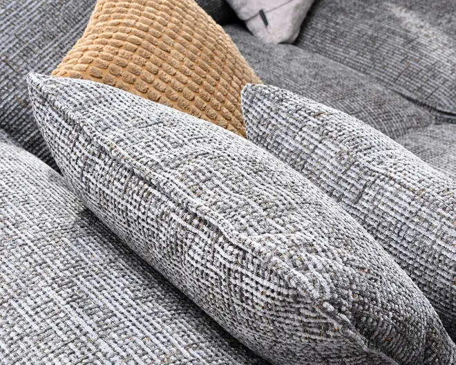 Nærbillede af teksturerede vævede pyntepuder i lysegrå og sandfarvet, arrangeret på en grå sofa.
