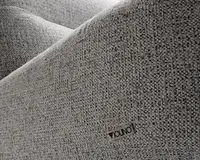 Nærbillede af et tekstureret, lysegråt stof med et diskret vævet mønster. Stoffet ser blødt og slidstærkt ud, velegnet til polstring. Et lille metalmærke med navnet 'VIOLINO' er fastgjort på overfladen og angiver producenten. Billedet fremhæver materialets kvalitet og detaljer.