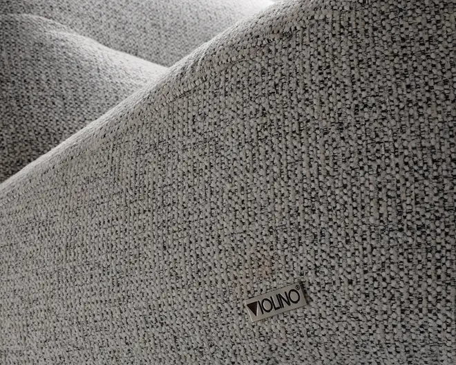 Nærbillede af et tekstureret, lysegråt stof med et diskret vævet mønster. Stoffet ser blødt og slidstærkt ud, velegnet til polstring. Et lille metalmærke med navnet 'VIOLINO' er fastgjort på overfladen og angiver producenten. Billedet fremhæver materialets kvalitet og detaljer.