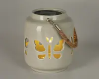 Keramisk lanterne med blank hvid overflade og udskæringer i form af sommerfugle, der lader varmt gult lys skinne igennem. Lanternen har et robust håndtag af reb, fastgjort med metalbeslag, hvilket gør den nem at bære eller hænge op. Øverst på lanternen er der et solpanel til opladning, hvilket gør den velegnet til udendørs brug.