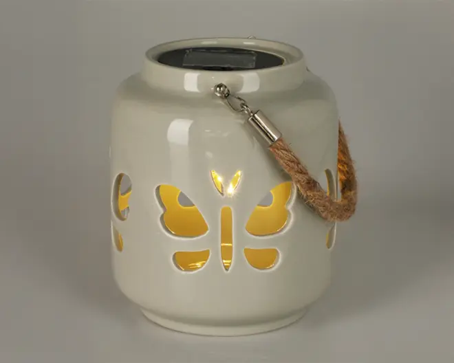 Keramisk lanterne med blank hvid overflade og udskæringer i form af sommerfugle, der lader varmt gult lys skinne igennem. Lanternen har et robust håndtag af reb, fastgjort med metalbeslag, hvilket gør den nem at bære eller hænge op. Øverst på lanternen er der et solpanel til opladning, hvilket gør den velegnet til udendørs brug.