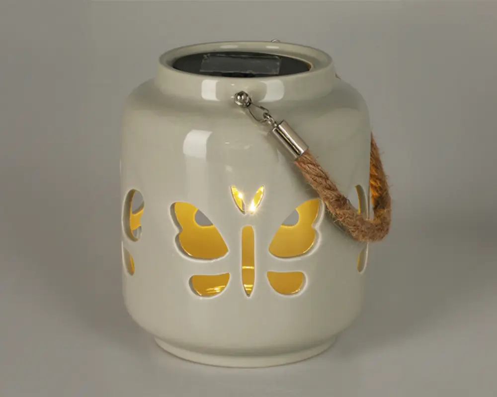 Keramisk lanterne med blank hvid overflade og udskæringer i form af sommerfugle, der lader varmt gult lys skinne igennem. Lanternen har et robust håndtag af reb, fastgjort med metalbeslag, hvilket gør den nem at bære eller hænge op. Øverst på lanternen er der et solpanel til opladning, hvilket gør den velegnet til udendørs brug.