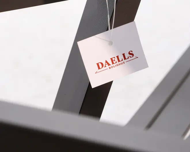 Nærbillede af et hvidt, rektangulært mærke, der er fastgjort til en mørk metalramme med en tynd snor. Mærket har mærkenavnet 'DAELLS BOLIGHUS' i fede, røde bogstaver med en enkel understregning. Metalrammen har en mat overflade og rene, vinklede linjer.