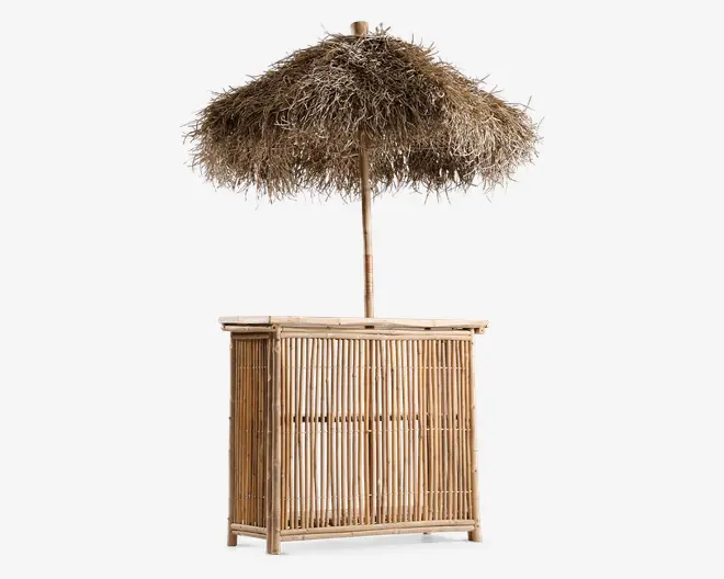 Fritstående bardisk lavet af naturlig bambus med lodrette lameller, udstyret med en bred bordplade og en stor, fastmonteret parasol med stråtag for skygge. Parasollet er lavet af tørret græs eller palmeblade, hvilket giver et tropisk og rustikt udseende. Baren har en åben bagside for nem adgang og opbevaring, og det samlede design er let og velegnet til udendørs brug.