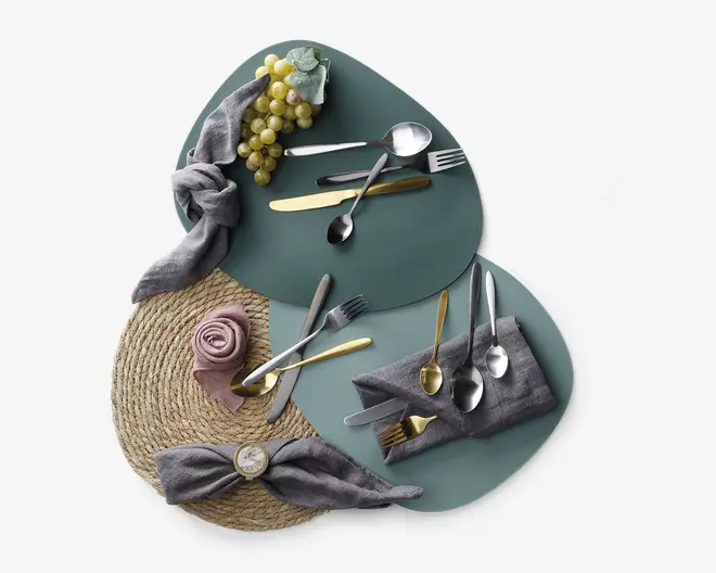 Et sæt bestik med en blanding af guld- og sølvfinish, herunder gafler, knive og skeer. Bestikket er arrangeret på to teal-farvede, organisk formede dækkeservietter, med grå stofservietter foldet på forskellige måder og en rund dækkeserviet af flet. Dekorative elementer inkluderer en klase grønne druer og en lille mintgrøn serviet. Præsentationen er elegant og moderne, velegnet til et stilfuldt bordopdækning.