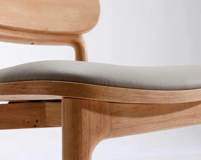 Nærbillede af en stol med en glat ramme af lyst træ og afrundede kanter. Sædet er polstret og betrukket med et blødt, lysegråt materiale, der giver komfort. Ryglænet er også lavet af det samme lyse træ og har et let buet design for ergonomisk støtte. Håndværket fremhæver rene linjer og et minimalistisk udtryk.