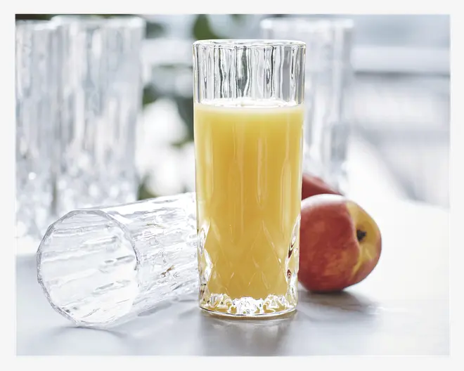 Højt, klart glas med en tekstureret, facetslebet overflade og lodrette riller, fyldt med appelsinjuice. Glasset har en tyk, solid bund og et moderne, elegant design. Et identisk tomt glas ligger på siden ved siden af, og to æbler er placeret ved glassene.