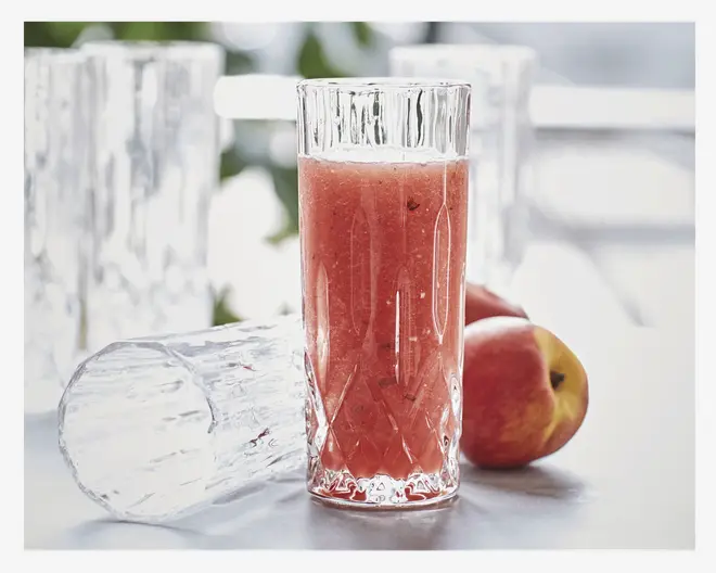 Højt, klart glas med facetslebet diamantmønster, fyldt med en rød frugtdrik. Glasset har en tyk bund og lodrette riller langs siderne, hvilket giver det et tekstureret og elegant udseende. I baggrunden ses et identisk glas, der ligger ned, samt to friske æbler, hvilket antyder, at drikken er lavet af æbler.