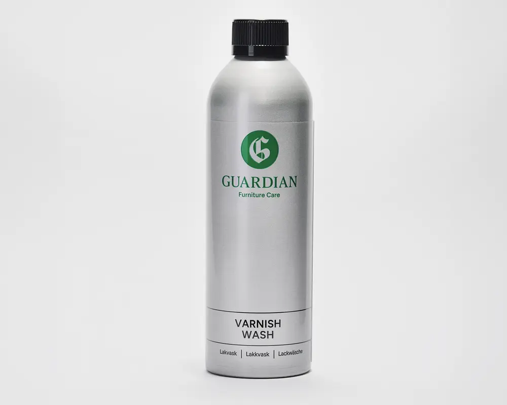 En høj, cylindrisk sølvfarvet flaske med sort skruelåg. Etiket med grønt 'G'-logo og teksten 'Guardian Furniture Care' i grøn. Under dette står der 'Varnish Wash' med sort tekst. Flasken har et enkelt og minimalistisk design med yderligere tekst nederst på etiketten på flere sprog.