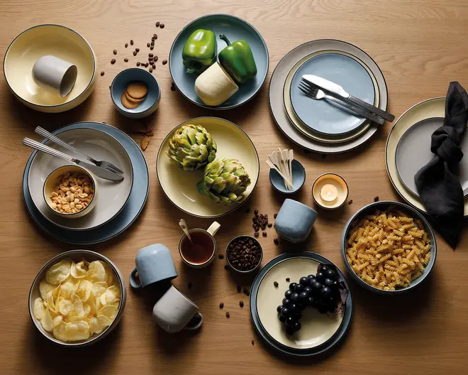 Et sæt koordineret keramikservice i afdæmpede blå, creme og grå nuancer, herunder tallerkener, skåle, krus og kopper. Sættet har et moderne, minimalistisk design med glatte, afrundede kanter og mat finish. Delene vises i forskellige størrelser, velegnet til servering af hovedretter, tilbehør og drikkevarer. Service sættet er vist med forskellig mad og bestik, hvilket fremhæver dets alsidighed til forskellige måltider.