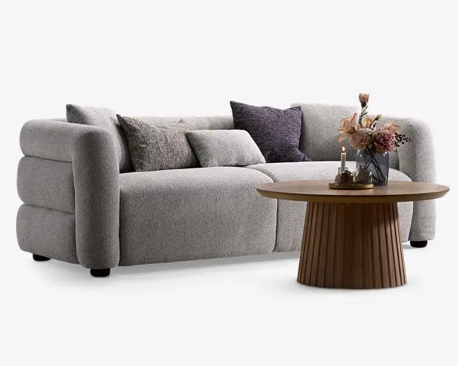 Lysegrå stofbetrukket 3-personers sofa med afrundede armlæn og flere teksturerede pyntepuder, kombineret med et rundt træ sofabord med riflet sokkel og blomsterdekoration.