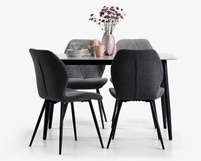 Et moderne spisebordssæt med et rektangulært bord med hvid bordplade og sorte ben, omgivet af fire polstrede stole i mørkegråt stof. Stolene har buet ryglæn med diskrete lodrette syninger og slanke sorte metalben. Bordet er pyntet med en lyserød vase med friske blomster, stablede tallerkener, skåle og krus i pastelfarver, hvilket skaber en hyggelig og indbydende stemning.