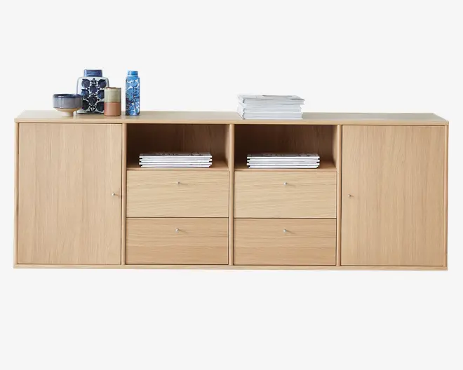 Et rektangulært skab i lyst egetræ med glat overflade. Skabet har to lukkede skabe i hver ende med enkle runde metalgreb, to åbne hylder i midten med stakke af magasiner, samt fire rummelige skuffer under hylderne med matchende metalgreb. Øverst står forskellige genstande, herunder stakke af bøger, keramiske skåle, kopper og dekorative vaser i blå og neutrale nuancer.