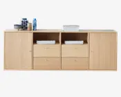 Et rektangulært skab i lyst egetræ med glat overflade. Skabet har to lukkede skabe i hver ende med enkle runde metalgreb, to åbne hylder i midten med stakke af magasiner, samt fire rummelige skuffer under hylderne med matchende metalgreb. Øverst står forskellige genstande, herunder stakke af bøger, keramiske skåle, kopper og dekorative vaser i blå og neutrale nuancer.