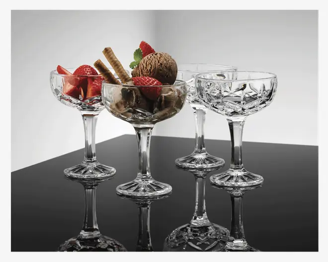 Sæt med fire klare dessertglas med facetslebet, diamantmønstret skål og en solid, bred fod. Glassene har en bred og lav skål, der egner sig til servering af is eller frugt. Ét glas er fyldt med chokoladeis, friske jordbær og vaffelruller, mens de andre er tomme, hvilket fremhæver det elegante design og den reflekterende overflade.